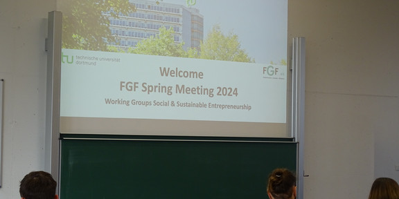 Ein Bild der Wilkommens-Präsentation zum Spring Meeting. Man sieht die erste Folie an die Wand projeziert und Leute von hinten, die davor sitzen.